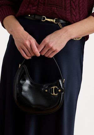 Melnas ādas rokassomu ar spīguļojošu apdari, izliekta forma, zelta aksesuāri un īsa roktura, kas turēta rokās pret tumšu apģērbu.