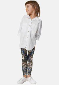 Fehér, gombos ing, két mellzsebbel, színes virágmintás leggings-szel, amelyen madarak láthatók, és fehér tornacipővel párosítva.