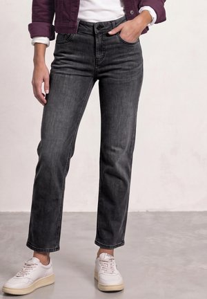 Personne portant un jean noir droit, des baskets blanches, une chemise blanche et une veste bordeaux, debout avec une main dans la poche.