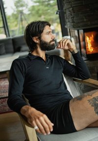 Sort langærmet top med zip-krave, afslappet pasform, kombineret med sorte shorts. Sidder i en træstol ved en stenvænge.