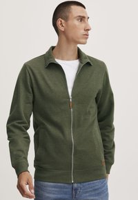 Grønn zip-up jakke laget av et mykt stoff, med ståkrage, sidelommer og ribbestrikket nedkant. Glatt tekstur uten mønstre.