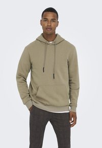 Light olive green hoodie with a front pocket, drawstring hood, and ribbed cuffs, paired with patterned dark trousers.