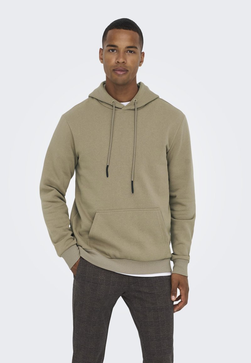 Light olive green hoodie with a front pocket, drawstring hood, and ribbed cuffs, paired with patterned dark trousers.