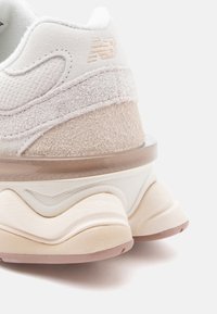 Beige idrettssko med teksturert semsket skinn og nettingpaneler, med en kraftig, bølgete såle og gjennomsiktige detaljer. Diskret merkevare på hælen.