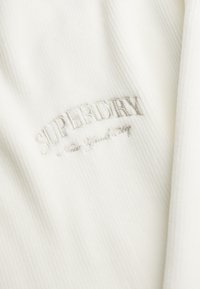 Maglione in tessuto a coste bianco con logo ricamato "SUPERDRY" e dettaglio "New York City" in filo argentato. Texture morbida, design casual.