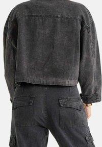 Persona che indossa un giubbotto di jeans nero sbiadito, corto, e pantaloni cargo abbinati con tasche a patta, mostrata di spalle.