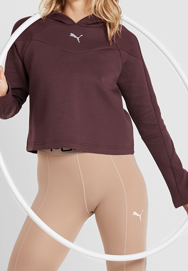 Bordeauxfarbener Kapuzenpullover mit cropped Design und weißem Logo, kombiniert mit beigen Leggings, die kontrastierende Nähte und Logo-Details aufweisen.
