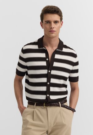 Junger Mann steht mit den Händen in den Taschen, trägt ein schwarz-weiß gestreiftes Kurzarmhemd mit Knöpfen und beige Hose mit einem geflochtenen braunen Gürtel.