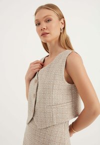 Beige tweed väst med en boxig design, breda ärmhål och två framknappar. Har en subtilt texturerad mönster och släta fållkanter.