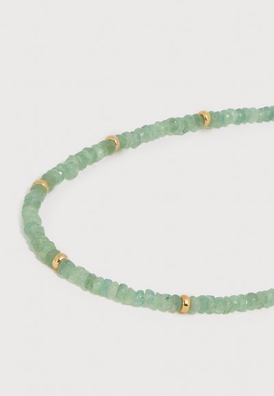 Κοντινό πλάνο ενός κολιέ με διάφανες πράσινες χάντρες και μικρές χρυσές μεσαίες χάντρες, τοποθετημένο σε κυκλικό μοτίβο σε λευκό φόντο.