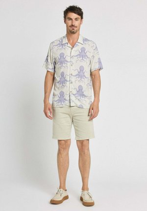Homme debout portant une chemise crème à manches courtes avec un imprimé pieuvre violet, un short beige et des chaussures beige clair sur un fond blanc uni.