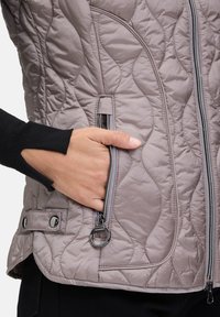 Gequilted grijze vest met een glanzende afwerking, ritszakken en accenten van zilverkleurig beslag. Inclusief een zwarte ribgebreide manchetdetail.