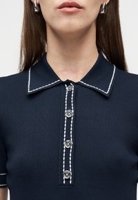 Κοντινό πλάνο ενός ατόμου που φοράει πουκάμισο σκούρου μπλε με λευκή ραφή και τέσσερα ασημένια κουμπιά στο μπροστινό μέρος, καθώς και ασημένια σκουλαρίκια κρίκους.