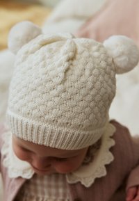 Cappello bianco lavorato a maglia con motivo a trama, caratterizzato da due soffici pompon sulla sommità e una fascia a coste alla base.