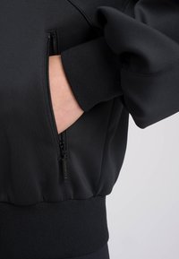 Felpa nera con tasca con zip, polsini a coste e design leggermente aderente con texture di tessuto opaco e liscia.