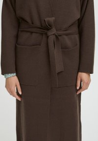 Persona che indossa un cardigan lavorato a maglia marrone con tasche frontali e una cintura annodata in vita, e una camicia con maniche arricciate di un blu chiaro sotto.