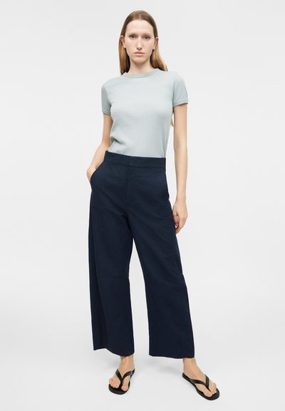 Γυναίκα με ίσια μαλλιά που φοράει γαλάζιο κοντομάνικο μπλουζάκι, φαρδιά μπλε σκούρα παντελόνια και μαύρα σανδάλια, στέκεται με το ένα χέρι στη τσέπη.