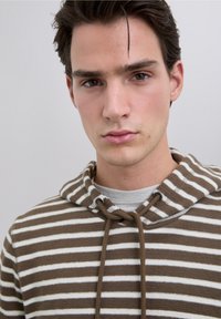 Sweat à capuche rayé marron et blanc avec une capuche à cordon et une texture douce, superposé sur un t-shirt gris à encolure ronde.