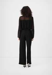 Femme aux cheveux bouclés portant un haut transparent noir à manches longues et un pantalon noir large, se tenant face à un fond blanc.