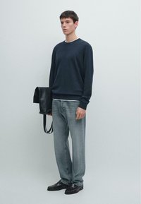 Granatowy sweter z długim rękawem, szare jeansy i czarne skórzane buty. Model trzyma czarną skórzaną torbę. Prosty, minimalistyczny styl.