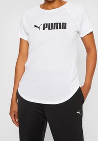 Valkoinen PUMA-t-paita, jossa on lyhyet hihat, pyöreä pääntie ja musta logo. Yhdistetty mustiin urheiluhousuihin, joissa on pieni valkoinen logo.