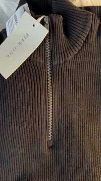 Mørk ribbet zip-up sweater med høj krave og en metallynlås. Inkluderer et mærkat med brandnavn og forhandlerinformation.