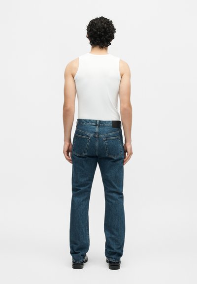 Άντρας με σγουρά μαλλιά που φοράει λευκό αμάνικο και μπλε τζιν, στέκεται γυρισμένος πλάτη σε απλό λευκό φόντο.