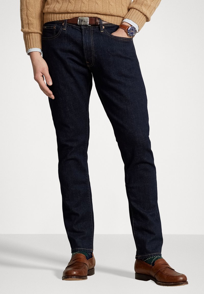 Blugi slim-fit de culoare albastru închis, cu cusături vizibile și buzunare, purtați cu mocasini maro și un pulover tricotat bej.