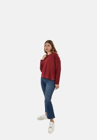 Maglia a maniche lunghe color bordeaux, texture a coste, vestibilità rilassata; abbinata a jeans a zampa d'elefante blu e sneakers bianche, in piedi contro uno sfondo semplice.
