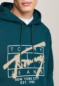 Sudadera de algodón en color verde azulado con un diseño de logo blanco y efecto de spray. Incluye cordones ajustables y una textura suave.
