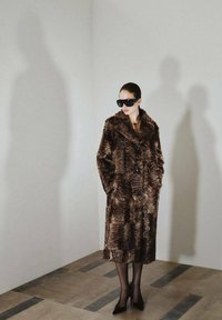 Cappotto in pelliccia sintetica marrone con un motivo a onde testurizzato, colletto ampio e due tasche anteriori, indossato con occhiali da sole neri e scarpe con tacco.