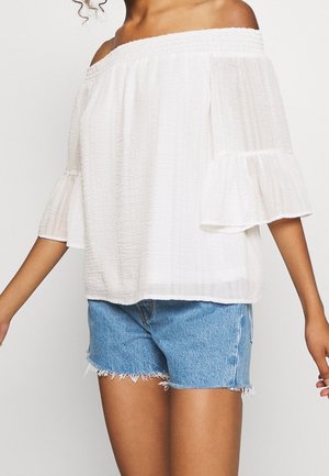 Witte top met blote schouders, korte uitlopende mouwen, getextureerde stof en een gerimpelde halslijn, gecombineerd met denim shorts met rafelige rand.
