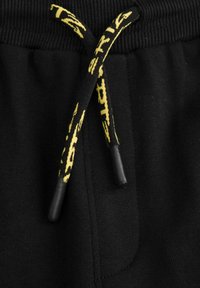 Czarne spodnie sportowe z teksturowanym pasem i sznurkiem ściągającym z żółtymi akcentami i czarnymi końcówkami. Zawierają boczną kieszeń.