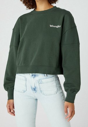 Kobieta w ciemnozielonej krótkiej bluzie Wrangler z prążkowanymi mankietami oraz jasnoniebieskich, wysokich dżinsach z przednimi kieszeniami.