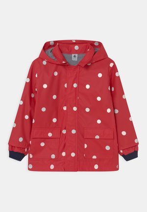 Rode kinderregenjas met witte stippen, capuchon, voorzakken met klep en marineblauwe boordjes, gestreepte voering zichtbaar.