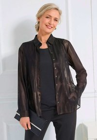 Glanzende vrouw met blond haar in een lage bun draagt een doorschijnende zwarte button-up blouse over een zwarte top, terwijl ze een zwarte clutch vasthoudt in een heldere kamer.