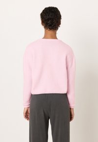 Osoba s tmavými kudrnatými vlasy sepnutými do drdolu, oblečená v světle růžovém svetru a tmavě šedých kalhotách, stojící zády k nám před prostým pozadím.