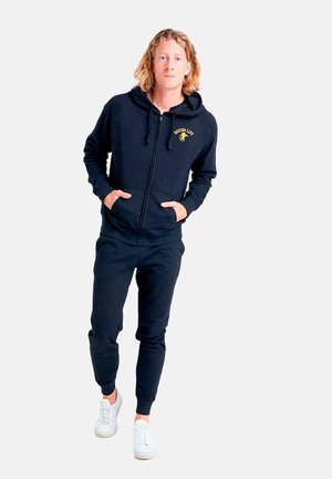 Jeune homme aux cheveux blonds portant un sweat à capuche noir et un pantalon avec des baskets blanches, debout les mains dans les poches devant un fond blanc.