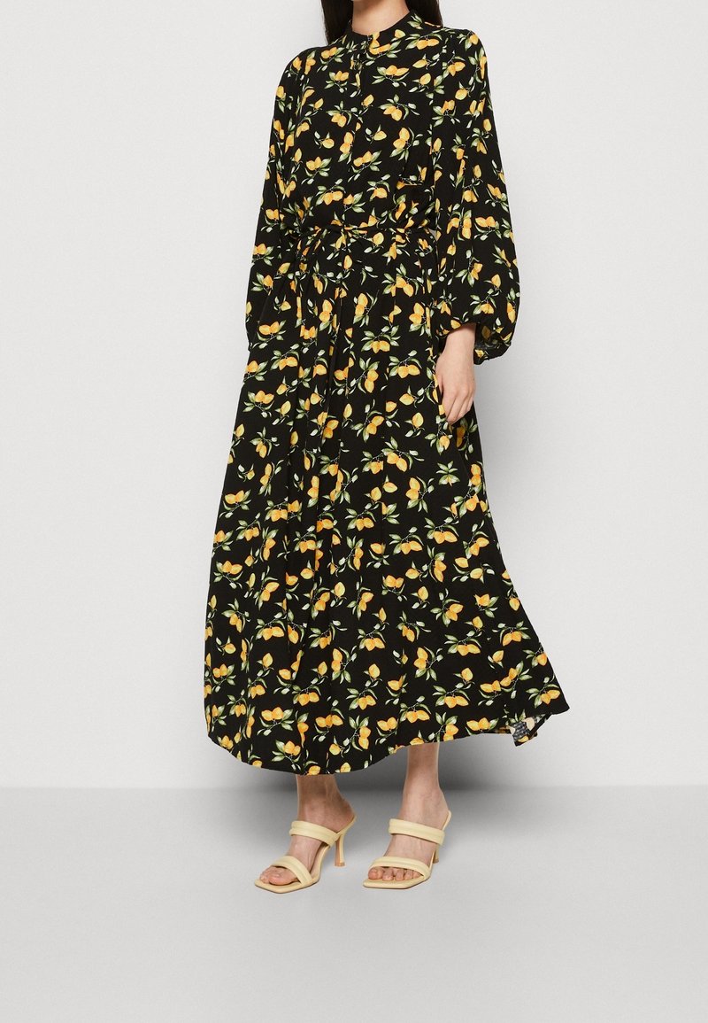 Donna che indossa un vestito midi nero con maniche lunghe e una stampa floreale di frutta arancione, abbinato a sandali con tacco beige.