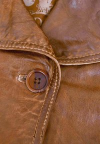 Braune Lederjacke mit glatter Textur, die einen auffälligen runden Knopf und gestickte Details entlang des Kragens und der Kanten aufweist.