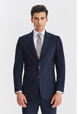 Granatowa marynarka z wyciętymi klapami, dwoma guzikami i bocznymi kieszeniami; zestawiona z białą koszulą i wzorzystym krawatem. Dopasowany fason z gładkiej tkaniny.