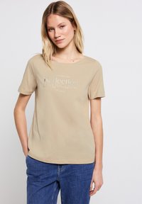 Beige tričko s krátkým rukávem vyrobené z měkké tkaniny s kulatým výstřihem. Na středu je potisk s reflexním stříbrným textem. V kombinaci s modrými džíny.