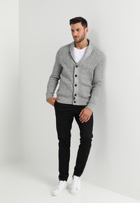 Grå strikket cardigan med ribbet tekstur, med sjalkrage, knappelukking foran og to frontlommer, i kombinasjon med svarte bukser og hvite joggesko.