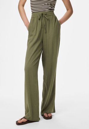 Άτομο με φαρδιά παντελόνια λαδί με κορδόνι, ριγέ αμάνικο τοπ και καφέ πλατφόρμες, στέκεται με τα χέρια στις τσέπες.