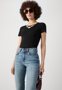 Svart kortermet topp med en kryssdesign i halsen, parret med lyseblå høytlivsjeans og en vinrød håndveske.
