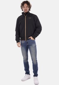 Schwarze Bomberjacke mit einem Frontreißverschluss und einem hohen Kragen. Mit einem goldenen Logo auf der Brust und gerippten Bündchen. Kombiniert mit blauen Jeans.