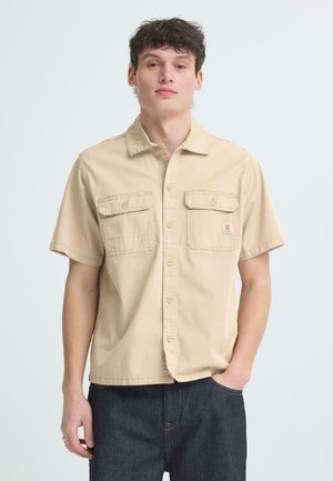 Unge mann iført beige kortermet skjorte med knapper og to brystlommer, og mørke jeans, stående med den ene hånden i lommen.