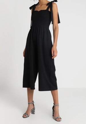 Femme portant une combinaison noire sans manches avec bretelles nouées aux épaules et pantalon large court, associée à des escarpins ouverts en cuir de serpent.