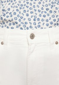 Calças de ganga brancas com uma cintura ajustada, apresentando elementos em metal e uma blusa com padrão floral em branco e azul a espreitar pela cintura.