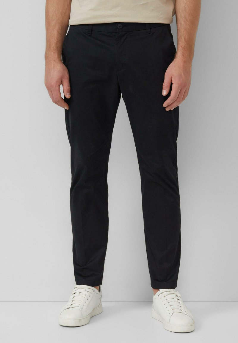 Man in slim-fit zwarte broek en witte sneakers, staand tegen een effen lichtgrijze achtergrond.